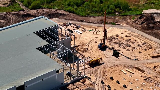 Aerial view of a large building frame under construction with a drilling rig and heavy machinery on a sandy site in Riga, Latvia.