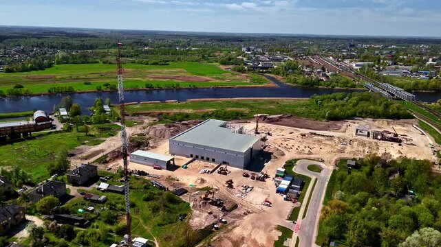 Aerial view of a large warehouse construction site with heavy machinery and a tall communications tower near a river and railway tracks in Riga, Latvia.
