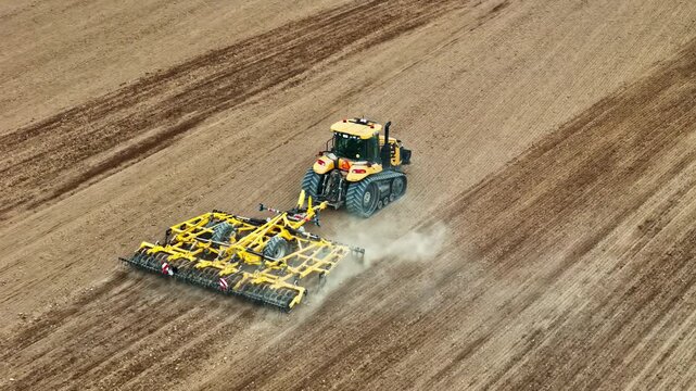 Aerial view of a yellow tractor pulling a large agricultural cultivator across a tilled brown field, creating dust in the rural landscape of Riga, Latvia.