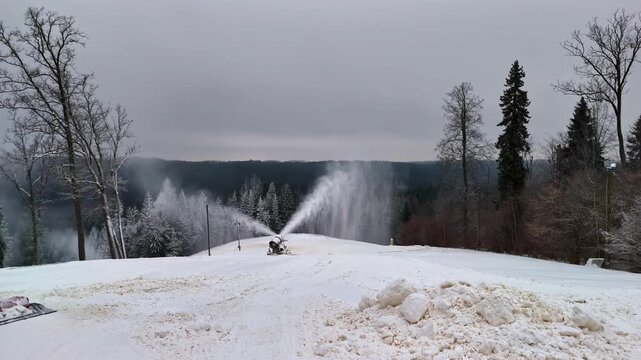 Aerial view of a snow cannon operating on a snowy slope with a snow groomer nearby and a forest background in Riga, Latvia.