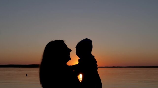 Happy mother holding her child and playing with him against beautiful sunset over lake or river. Tender and emotional silhouette of mother and kid bond and serene summer evening. Mom throws baby up