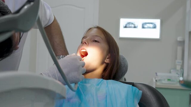 Pediatric dentist examining teeth of young girl sitting in dental chair.