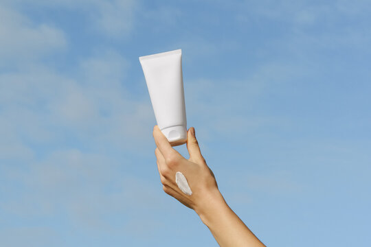 Woman's hand holds a blank white cosmetic tube against a blue sky. A swatch of cream shows its texture. Concept for sun protection, skincare, summer vacation, and beauty products mockup