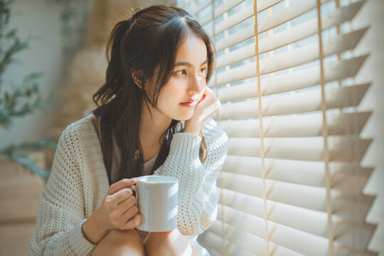 Young Asian woman holding coffee mug looking out window blind during morning sunlight, beautiful thoughtful female relaxing at home bedroom for cozy leisure lifestyle, mental health, wellness