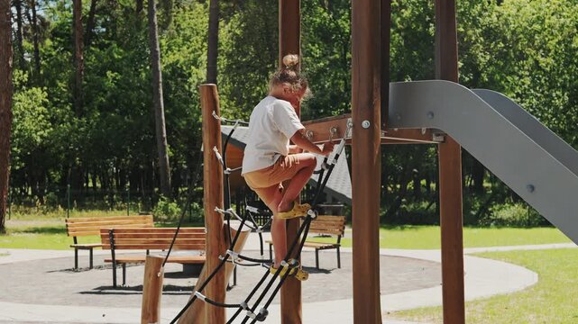 A child climbs on a playground structure and prepares to slide down. The park has green trees and sunny weather, creating a fun atmosphere for play