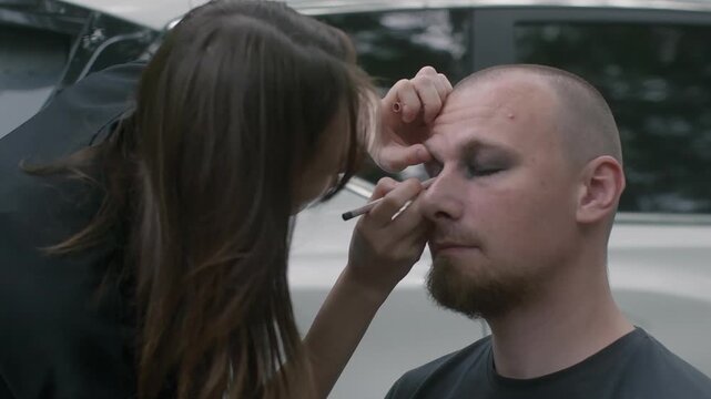 Professional makeup artist applies dark eyeshadow, eyeliner to bearded man during outdoor beauty transformation session for alternative, rock, or theatrical look.