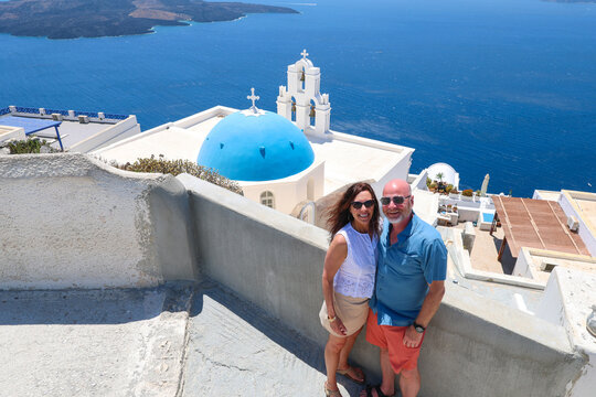 A couple stands before a blue‑domed church in Santorini, Greece, surrounded by whitewashed architecture and sweeping Aegean Sea views in a romantic Mediterranean setting.