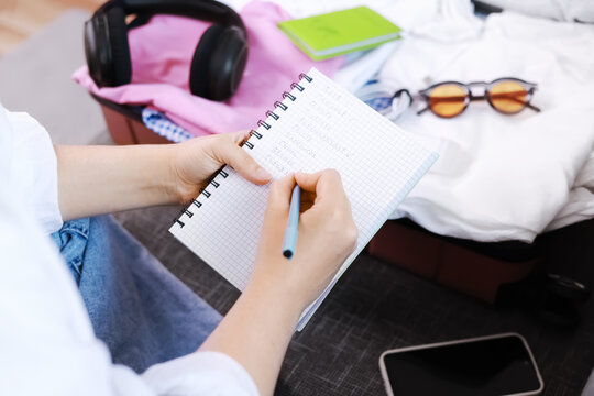 Woman writing a packing list in a notebook during travel and vacation preparation. Packed luggage, suitcase, and baggage for summer tourism trip.