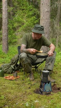 A military soldier in camouflage uniform and a green t shirt eating fresh hot meal directly from a frying pan while resting in a forest camp. Concept of army field rations, military training and comba