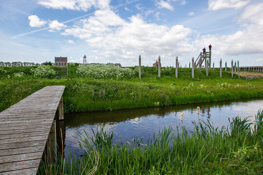 17 May 2015: View of a wooden bridge crossing a narrow canal with white wildflowers and a person standing on a wooden observation tower under a cloudy sky.