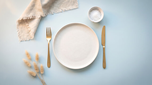 Flat lay of empty ceramic plate with gold cutlery on blue background and linen napkin with bowl of salt and dried rabbit tail grass for elegant minimalist table decoration.