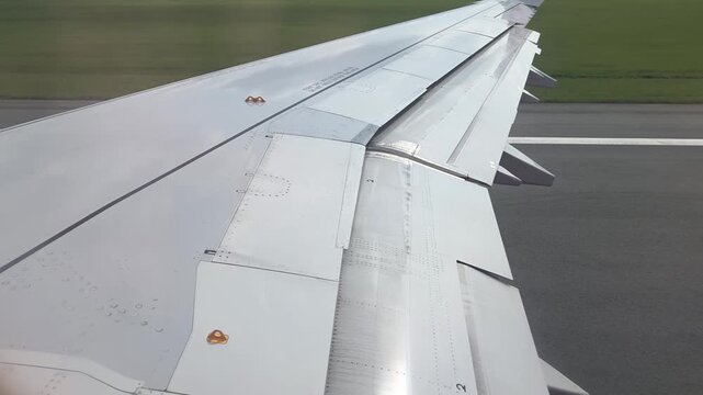 Aircraft wing with flaps extended during landing approach over green grass runway, showcasing aerodynamic surfaces and landing gear preparation for touchdown