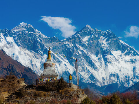 Aerial view of a Buddhist stupa with colorful prayer flags and a man standing on a stone platform against the backdrop of the snow-capped Mount Everest peaks in Nepal.