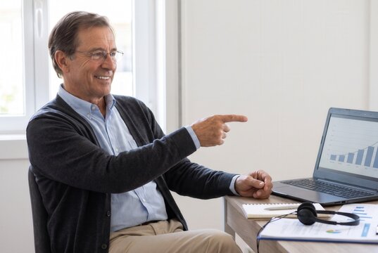 Senior man smiling, sitting at desk pointing at a laptop screen with a business growth chart