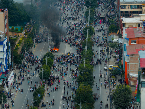 Aerial view of a large crowd of people gathered on a city street with a vehicle engulfed in flames and thick black smoke rising, surrounded by urban buildings and trees in Nepal.