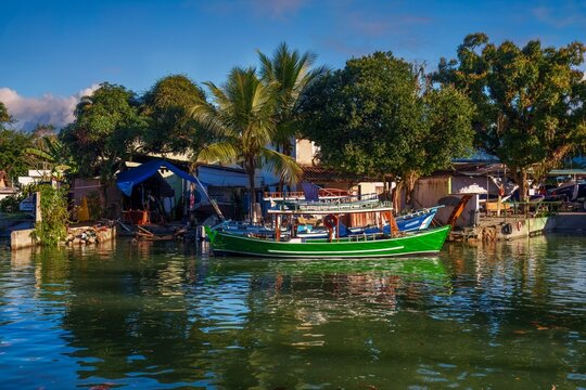 View of colorful boats on the Perequ&ecirc;-A&ccedil;u River canal bordered by tropical greenery and traditional houses, with Serra do Mar mountains under a blue sky in Paraty, Rio de Janeiro, Brazil.