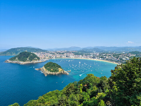 La Concha Bay and beach in San Sebastian, Spain, captured from Mount Igueldo. High-angle view of Santa Clara Island, turquoise water, and green hills under a clear blue sky.