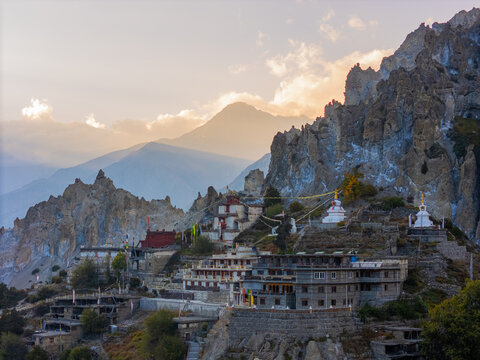 Aerial view of Braga Monastery nestled among rugged Himalayan peaks, featuring traditional stone architecture, white stupas, and fluttering prayer flags under the golden hour light in Nepal.