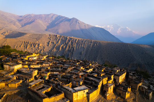 Aerial view of a traditional village with stone houses and flat roofs nestled in the rugged Himalayan mountain landscape during golden hour in Nepal.