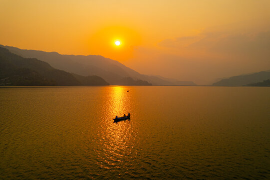 Aerial view of a small boat with people on a calm lake at sunset, with mountains in the background under a golden sky in Nepal.