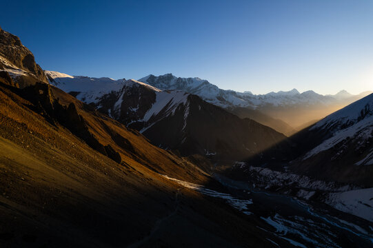 Aerial view of the Himalayan mountain range featuring snow-capped peaks, deep valleys, and dramatic golden hour light casting long shadows across the rugged terrain in Nepal.
