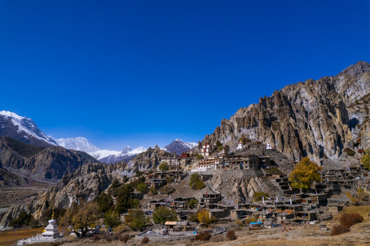 Aerial view of a traditional mountain village with stone houses and a monastery nestled among rugged rocky cliffs and snow-capped Himalayan peaks under a clear blue sky in Nepal.