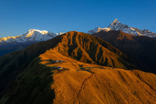 Aerial view of the golden-lit grassy ridges of the Himalayas with small mountain huts and snow-capped peaks in the background in Nepal.
