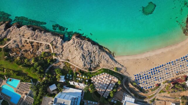 Aerial top-down view of Fig Tree Beach in Protaras, Cyprus. Clear turquoise water, sandy beach, rocky shoreline, resort pools, sunbeds and coastal paths create a beautiful Mediterranean summer holiday