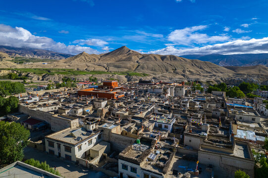 Aerial view of a traditional village in the Himalayas with flat-roofed stone houses, a red monastery, and colorful prayer flags set against arid mountain slopes in Nepal.