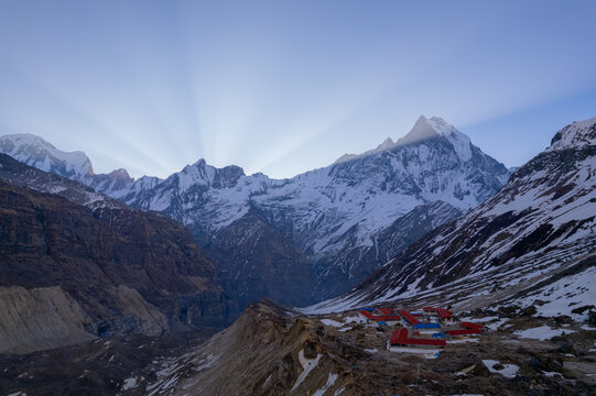 Aerial view of the snow-capped Machapuchare peak in the Himalayas with sun rays breaking over the summit and a base camp with red-roofed buildings in Nepal.