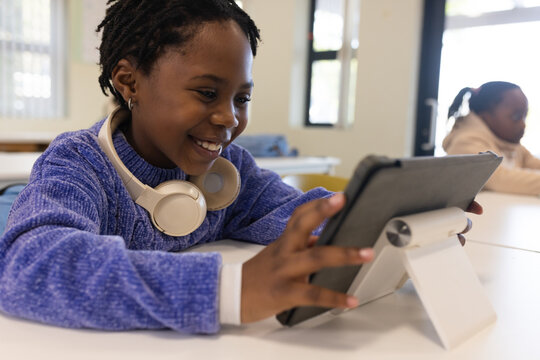 Female child classmates sitting at white table in classroom using tablet on white stand, copy space