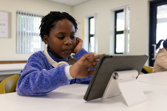 African youth girl sitting at desk in class using tablet on stand wearing headphones, blue sweater