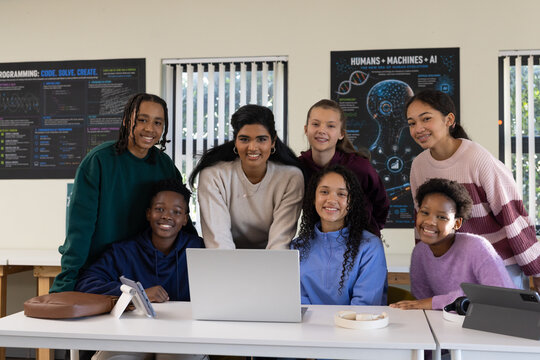 Diverse group of eight students leaning toward silver laptop, discussing project at classroom table