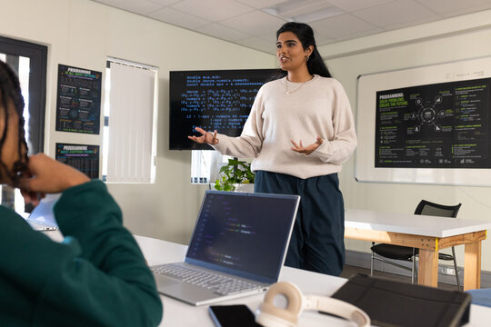 Female trainer in tan sweater gesturing while teaching in training room with laptop monitors poster