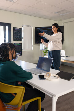 Woman teacher with teenage student presenting code on wall monitor, laptops, posters, copy space