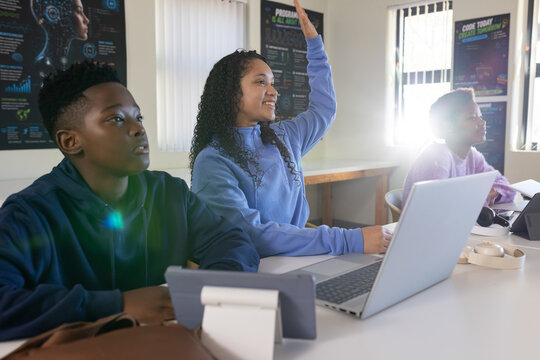 African youth classmates sitting at classroom table, female raising hand, using laptops and tablet