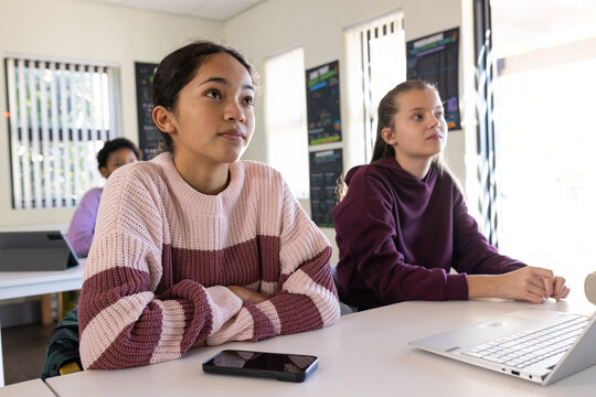 Diverse teenage female students sitting in class, in sweater, using phone and laptop, copy space