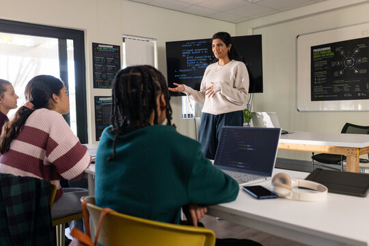 Indian-African American female instructor teaching coding to teenage coders in classroom on monitor