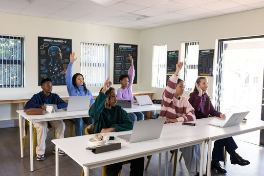 Diverse youth classmates raising hands and working on laptops in classroom with headphones
