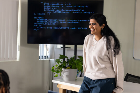 Adult Indian woman in tan top presenting code in office, monitor, plant, whiteboard, copy space