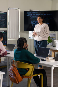 Indian woman instructor presenting code on monitor while teen students using laptops in classroom