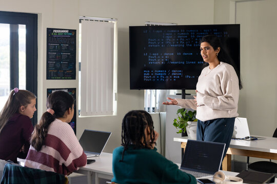 Indian instructor in light sweater presenting code to 3 teen female students in class with laptops