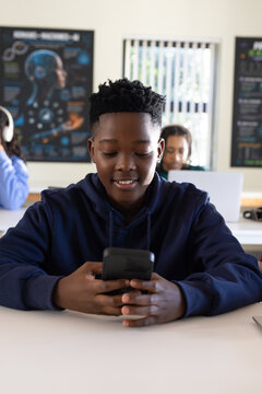 Classmates watching African American teen male student using smartphone at classroom desk, laptops