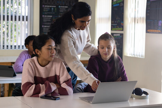 Teacher with two teenage female students leaning over laptop in classroom, wearing light sweater