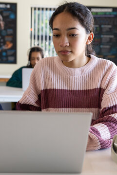 Teen classmates at desks with African American female wearing pink sweater typing on silver laptop