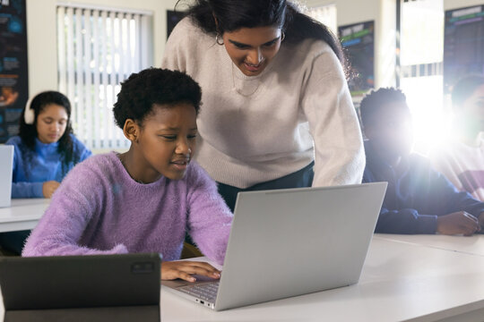 Indian teacher leaning over African teen wearing lavender sweater typing on laptop in classroom