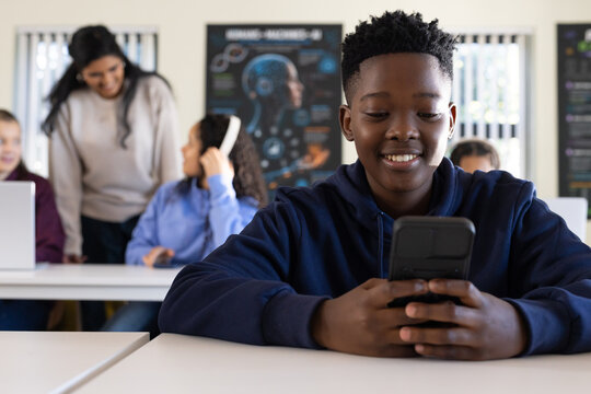 Diverse teen class working at desks with laptops, teen in navy hoodie using handheld device