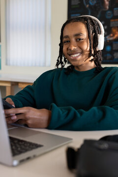 African American teenage male smiling while holding smartphone at desk with laptop and headset