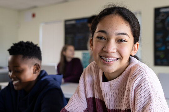 Teen classmates sitting at desks in classroom, smiling, wearing knit sweater, navy hoodie, braces