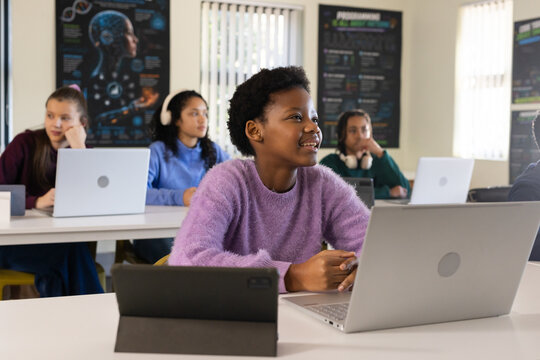 Diverse female teens studying in class, smiling pupil in purple sweater using silver laptop tablet